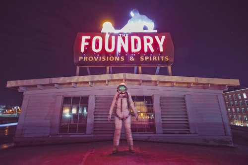 An astronaut stands on a rooftop in front of a neon sign reading "The Foundry Provisions & Spirits" at night.