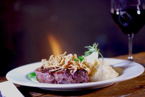 A plated steak with crispy onions, mashed potatoes, and a glass of red wine in the background.