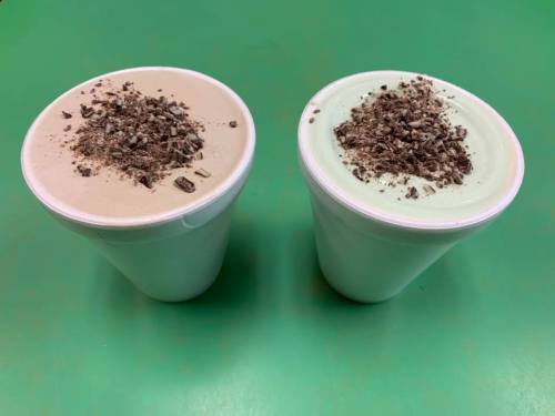 Two cups of ice cream topped with crushed chocolate, one light brown and one mint green, on a green surface.