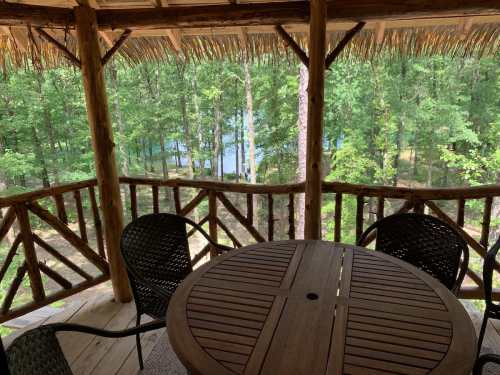 A wooden deck with a round table and chairs overlooks a serene forest and a body of water in the background.