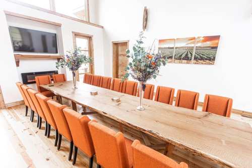 A spacious dining room with a long wooden table and orange chairs, decorated with floral centerpieces and a TV on the wall.