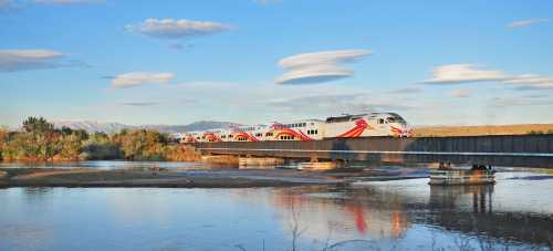 A train crosses a bridge over a river, surrounded by trees and mountains under a blue sky with clouds.