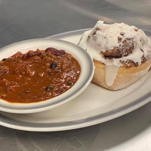 A plate with a bowl of chili and a cinnamon roll topped with icing.