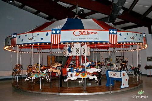 A vintage carousel with colorful horses, under a red, white, and blue striped canopy, in a spacious indoor setting.