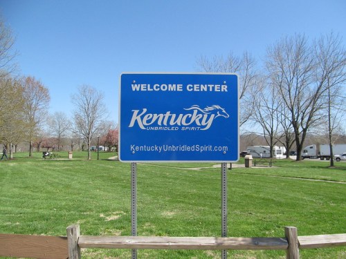 Blue welcome sign for Kentucky, featuring "Unbridled Spirit" and a website link, set in a grassy area with trees.