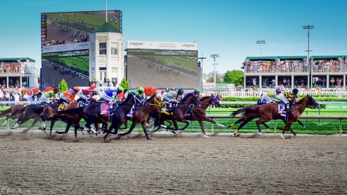 A lively horse race with multiple horses and jockeys competing on a dirt track, surrounded by spectators and large screens.