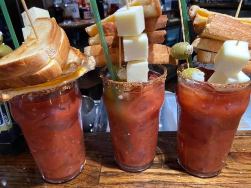 Three tall glasses of Bloody Marys topped with skewers of grilled cheese, bread, cheese cubes, and olives.