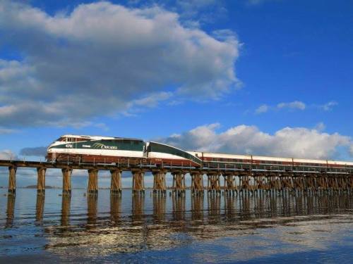 A train travels along a pier over calm water, with blue skies and clouds in the background.
