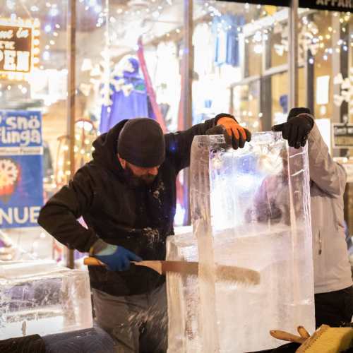 Two people sculpting ice blocks outdoors, surrounded by festive lights and decorations.