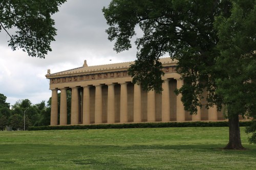 A large, classical-style building with columns, set against a cloudy sky and surrounded by green grass and trees.