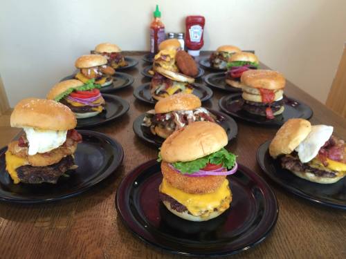 A variety of gourmet burgers on black plates, featuring toppings like cheese, bacon, and sauces, arranged on a wooden table.