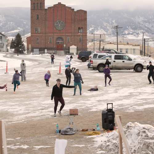 A group of people exercising outdoors in a snowy area, with a church and parked cars in the background.