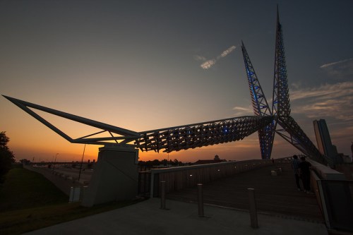 A modern sculpture resembling a large, abstract bird against a sunset sky, with city buildings in the background.