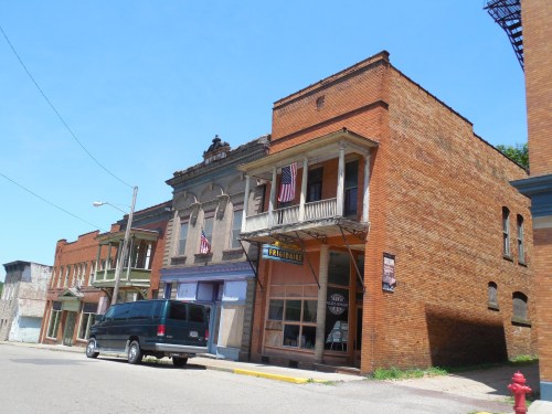 Abandoned brick buildings with American flags, a parked van, and a clear blue sky in a small town setting.