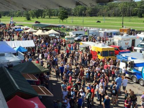 A bustling outdoor event with large crowds, food trucks, and tents set up in a sunny area.