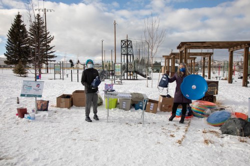 Two people in winter attire stand in a snowy park, surrounded by colorful equipment and supplies for outdoor activities.