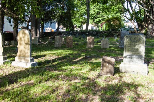 A peaceful cemetery with various gravestones surrounded by trees and grass under dappled sunlight.