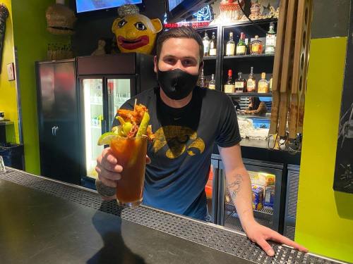 A bartender wearing a mask holds a large cocktail with garnishes at a vibrant bar.