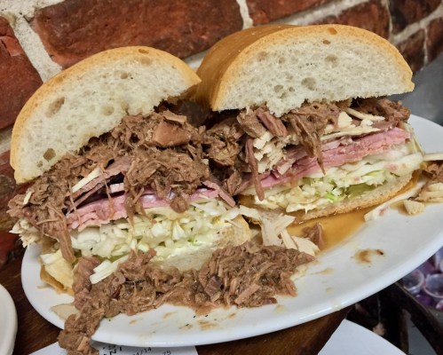 A hearty sandwich filled with shredded meat, ham, and cabbage, served on a soft bun, with juices spilling out.
