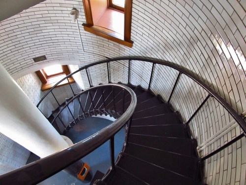 A spiral staircase with white tiled walls and wooden windows, viewed from above.