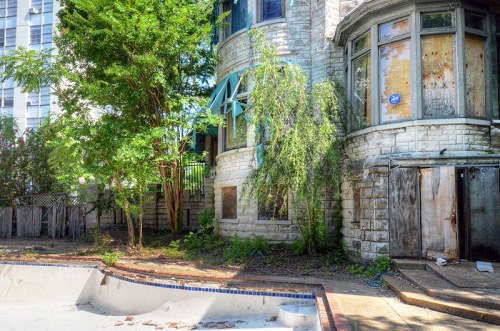 Abandoned building with boarded windows, overgrown vegetation, and a dry pool in the foreground.