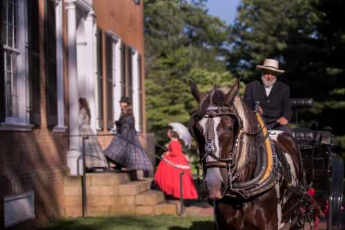 A man in a hat rides a horse near a historic building, while women in period dresses walk nearby.