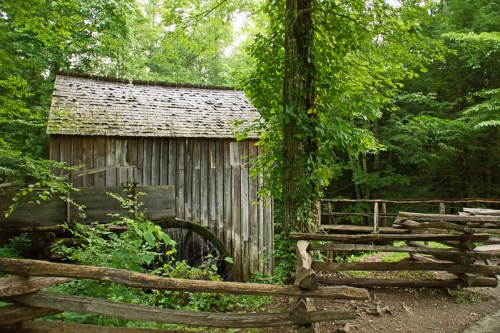 A rustic wooden mill surrounded by lush green trees and a wooden fence in a serene forest setting.