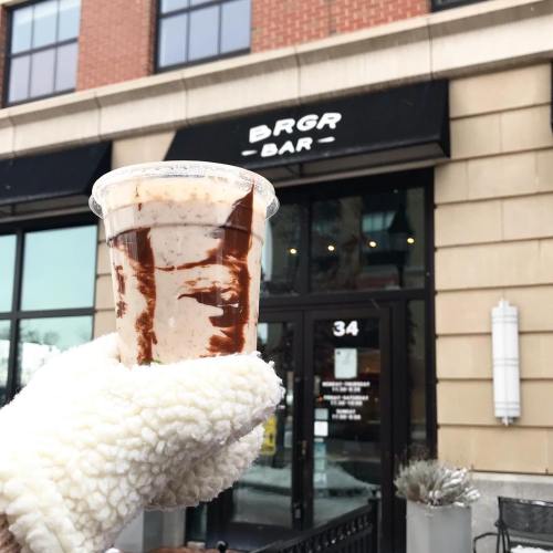 A hand in a fluffy white glove holds a dessert cup with chocolate swirls in front of a restaurant called BRGR Bar.