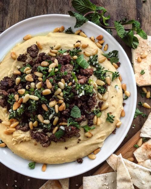 A plate of hummus topped with spiced ground meat, pine nuts, and fresh herbs, surrounded by pita bread.