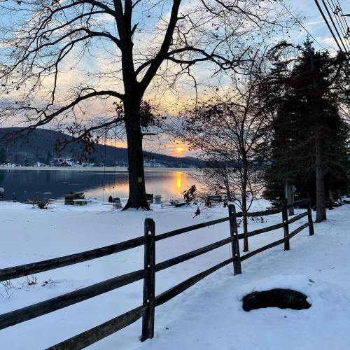 A serene winter scene with a sunset reflecting on a lake, framed by trees and a wooden fence. Snow covers the ground.
