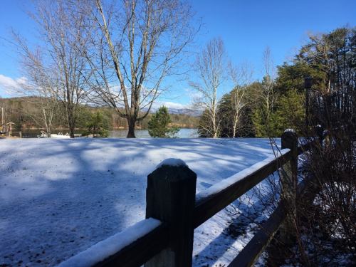 A snowy landscape with a wooden fence, bare trees, and a calm lake under a clear blue sky.