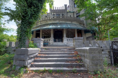 A stone staircase leads to a grand, overgrown entrance of an old mansion surrounded by trees and greenery.