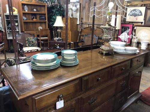 A wooden sideboard with stacked plates and a vintage lamp, surrounded by various furniture pieces in a cozy shop.