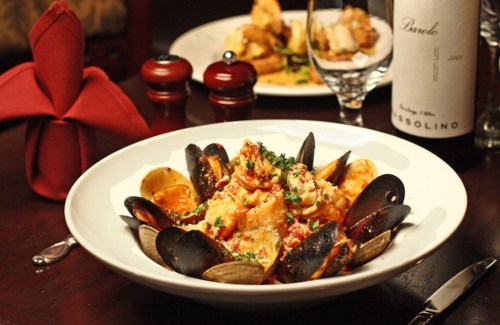 A bowl of pasta with mussels and clams, garnished with herbs, set on a dining table with red napkins and wine.