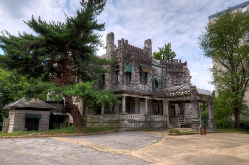 Abandoned stone mansion with gothic architecture, surrounded by trees and overgrown vegetation under a cloudy sky.