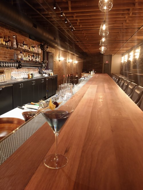 A sleek bar with a long wooden counter, dim lighting, and a blue cocktail in a glass.