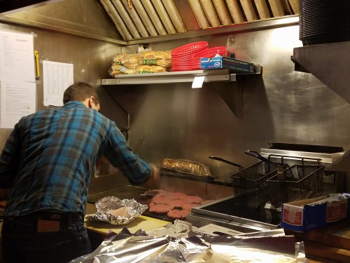 A cook in a plaid shirt grills burgers in a busy kitchen, with baskets and supplies visible in the background.