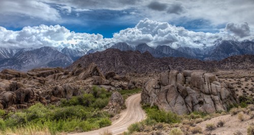 A winding dirt road leads through rocky terrain, with majestic mountains and dramatic clouds in the background.