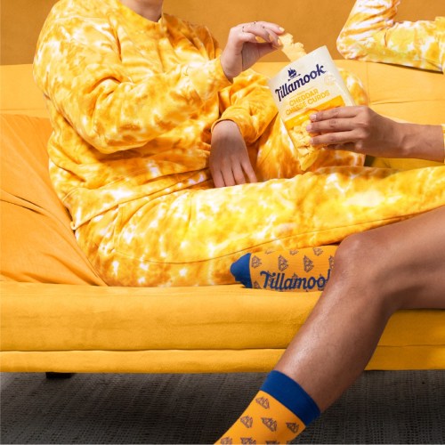 Two people in matching yellow outfits enjoy Tillamook cheese puffs while sitting on a bright yellow couch.
