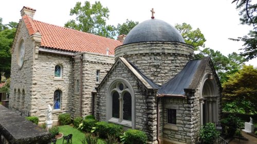 A stone chapel with a dome and cross, surrounded by greenery and trees under a clear blue sky.