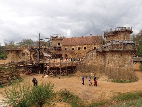 A medieval-style castle with wooden scaffolding, surrounded by visitors and greenery under a cloudy sky.