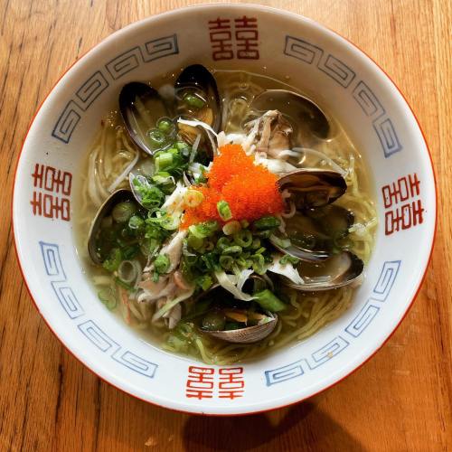 A bowl of ramen topped with clams, green onions, and orange fish roe, served in a decorative bowl on a wooden table.