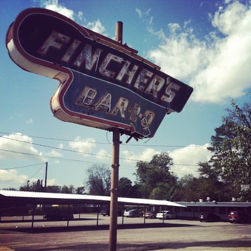 A vintage sign for Fincher's Bar-B-Q against a blue sky with clouds, featuring a retro design and surrounding trees.