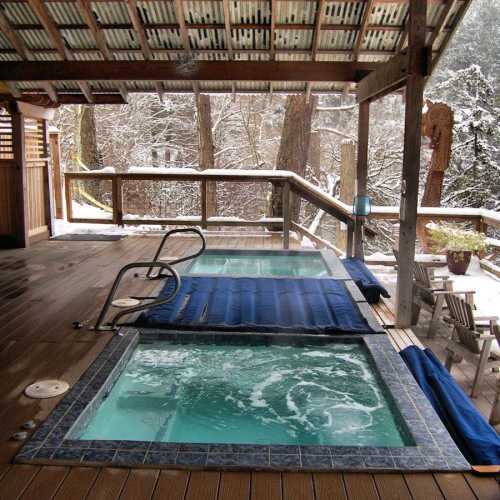 Two hot tubs on a wooden deck surrounded by snow-covered trees, creating a cozy winter retreat.