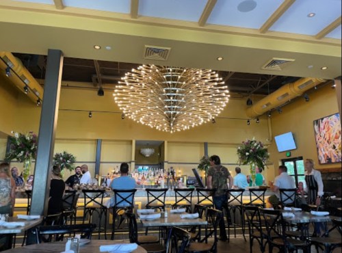 Interior of a restaurant featuring a large chandelier, bar area, and patrons seated at tables. Bright and modern decor.