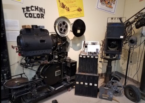 Vintage film equipment including a projector, cameras, and reels displayed in a museum setting.