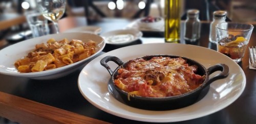 Two plates of pasta: one in a white bowl with rigatoni and sauce, the other in a cast iron dish, baked with cheese.