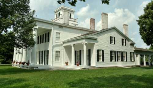 A large white historic house with columns, surrounded by green grass and trees under a partly cloudy sky.