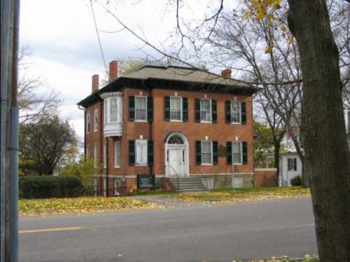 A historic brick house with green shutters, a white door, and a large front porch, surrounded by autumn foliage.