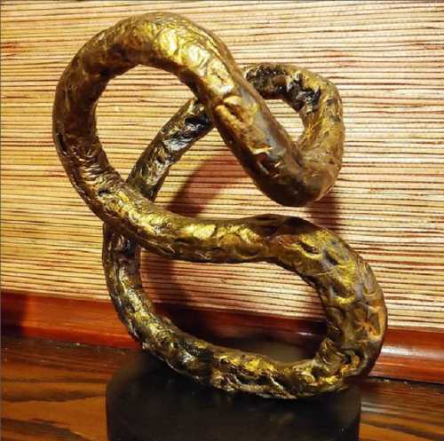 A twisted, abstract sculpture in gold and black, resting on a circular base against a textured background.